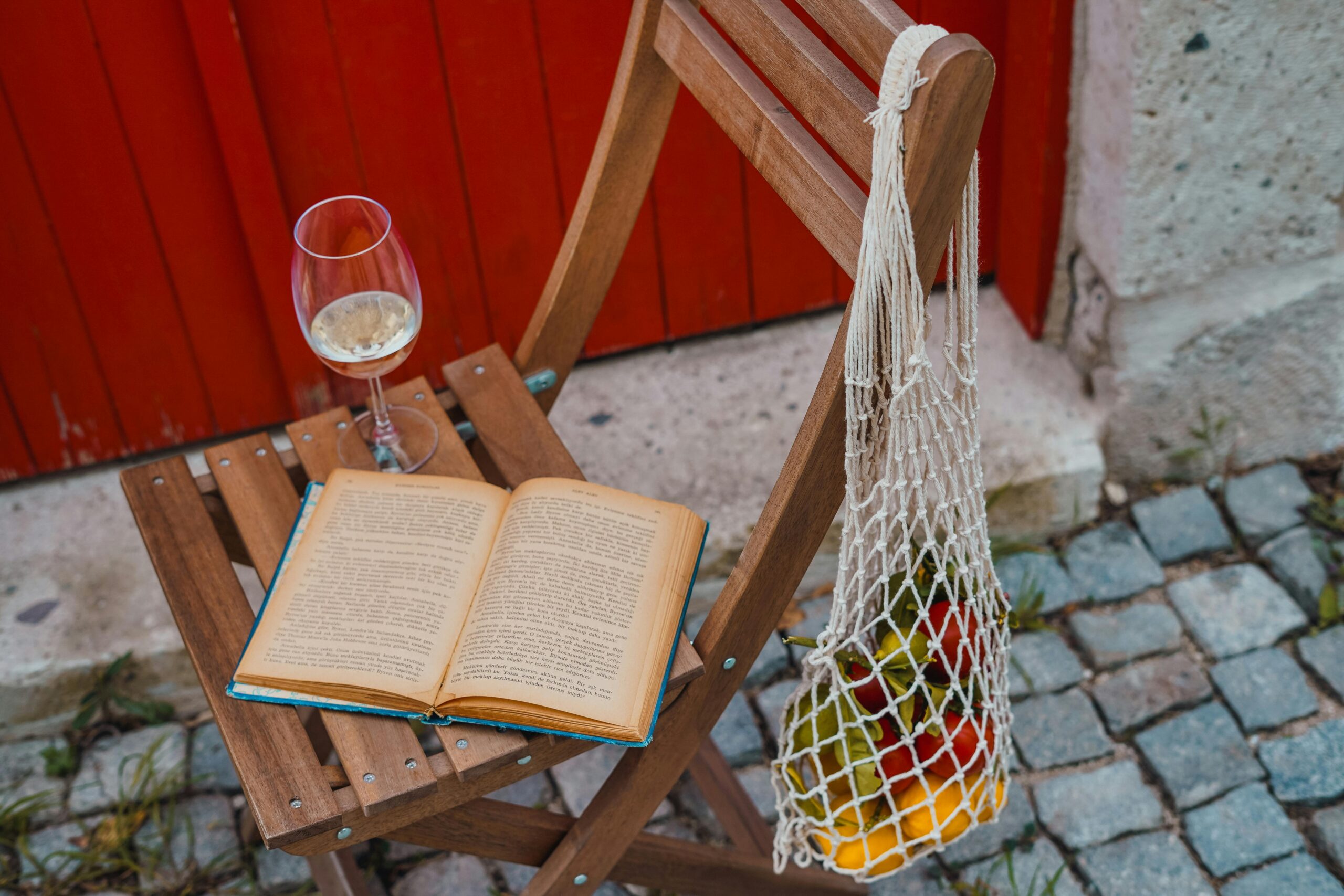 wooden chair with book and glass of white wine on the seat, a crochet bag with fruit hanging pn the side