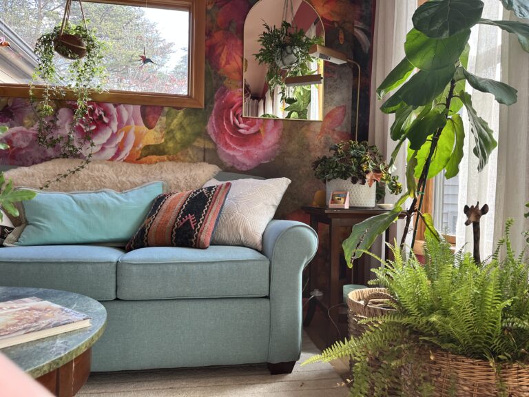 Photo of a teal colored sofa against a wall with flower wall paper and plants surrounding the sofa.